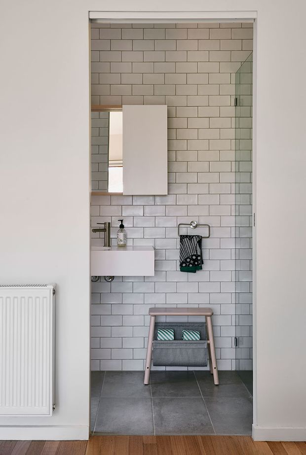 A compact contemporary bathroom viewed through an open doorway, with white subway-tile walls, dark grey floor tiles, a wall-mounted white vanity and basin, chrome tapware, a mirrored cabinet, and a glass shower screen at the right. A freestanding towel rail and small stool add practical storage. The bathroom opens from a larger room with timber flooring and a visible radiator.
