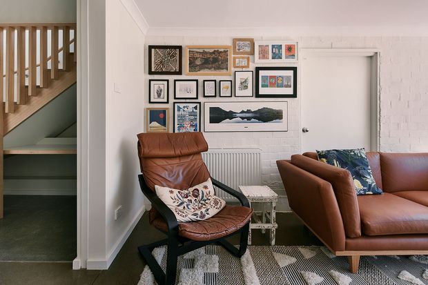 A warm, eclectic living room with a brown leather armchair and matching sofa arranged around a patterned rug. The white-painted brick and plaster walls display a curated gallery of framed artwork, while a white door and radiator sit behind the seating. A timber stair balustrade and stairway are visible through the opening on the left.