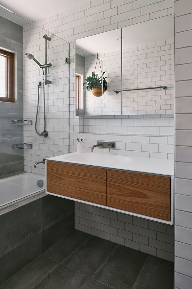 Contemporary bathroom with a wall-mounted timber vanity, white integrated basin and chrome wall-mounted tapware. White subway tiles cover the walls, while dark grey floor and lower shower-area tiles create contrast. A frameless mirrored cabinet, walk-in shower with rain showerhead, towel rail, and a hanging plant complete the clean, functional design.