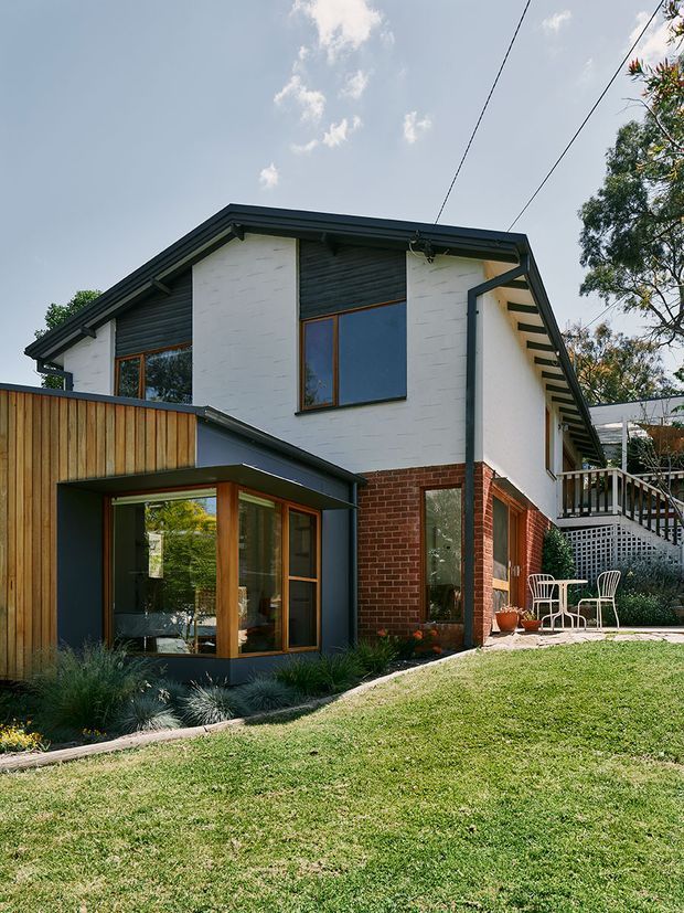 Exterior view of a two-storey contemporary residential house on a sloped garden site. The home combines white-painted brick upper walls, exposed red brick lower walls, dark horizontal cladding, dark roof framing, and warm timber-framed glazing. A projecting glazed extension sits to the left, while a raised outdoor terrace with white balustrades and a small paved seating area is visible to the right.