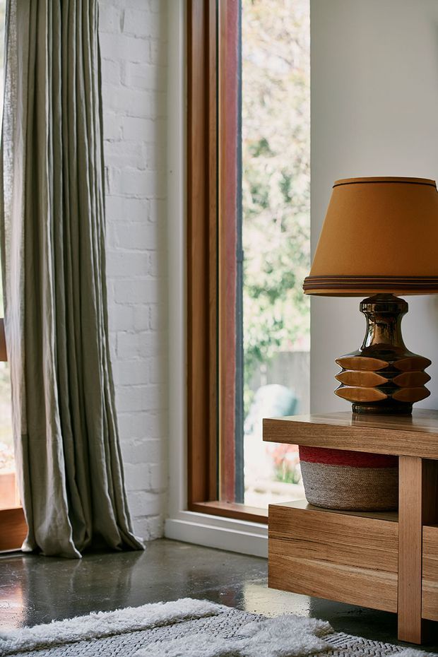Close-up interior detail of a warmly styled room featuring a tall timber-framed window, white-painted exposed brick wall, full-length olive-grey curtains, polished dark concrete floor, and a mid-century-inspired timber console with a ceramic table lamp and woven storage basket. A textured pale rug occupies the foreground, with leafy outdoor scenery visible through the window.