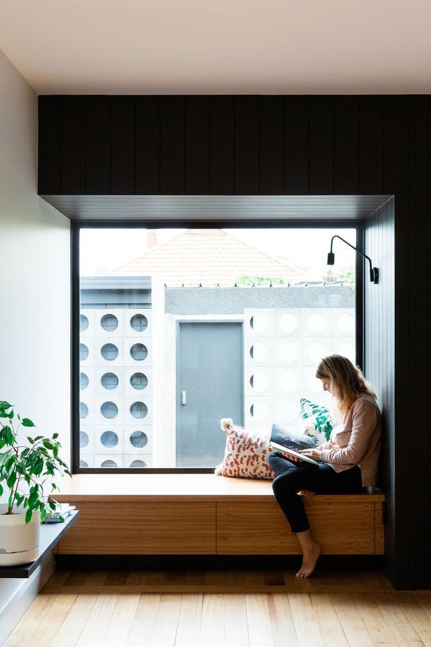 A bright, contemporary reading nook centered on a large window seat. Dark vertical timber cabinetry spans the upper wall and continues along the right side, contrasting with pale walls and light timber flooring. The built-in timber bench includes drawers and is styled with cushions, a small plant, and a seated person reading. Through the window, a modern exterior with perforated concrete block walls is visible.