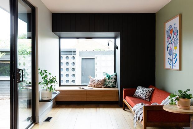 A compact, light-filled living room with pale timber flooring, a black vertical-panel feature wall and matching overhead joinery, large black-framed glazing, and a low timber window seat or cabinet. A mid-century-style rust-red sofa with patterned cushions sits against a muted green wall, accompanied by indoor plants and framed artwork. The room has a warm, contemporary mid-century character and overlooks an adjacent building through the window.