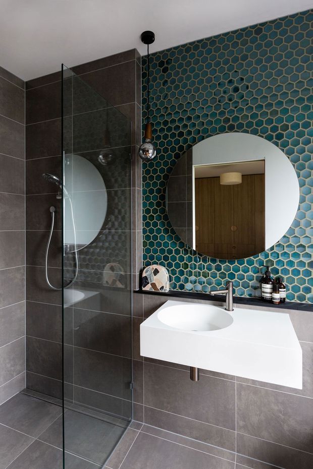 A contemporary bathroom with a frameless glass walk-in shower, dark large-format wall and floor tiles, and a wall-mounted white vanity with integrated basin. The focal wall is clad in blue-green hexagonal mosaic tiles with dark grout and a large round mirror. Chrome tapware and spherical pendant lights add reflective accents, while timber cabinetry is visible in the mirror reflection.