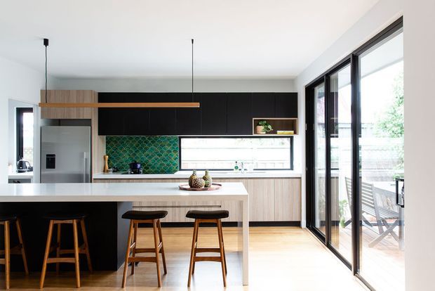 Bright contemporary kitchen with a large white island and timber bar stools, black lower and overhead cabinetry, light timber joinery, a green patterned tile splashback, and extensive black-framed glazed doors opening to an outdoor area. Minimal pendant lighting and a clean open-plan layout create an airy, refined space.