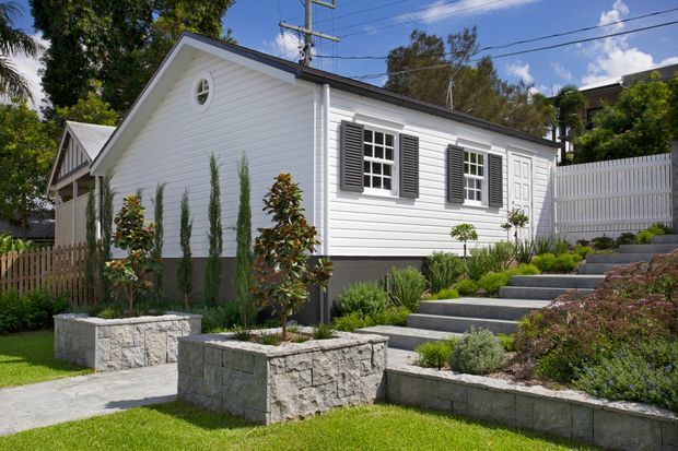 A small, single-storey white weatherboard house with a steep gabled roof, dark grey trim, multi-pane windows with dark shutters, and a circular attic window. The home is approached by broad concrete steps through neatly landscaped raised garden beds and low stone retaining walls, with a timber picket fence and neighbouring residential buildings visible around it.