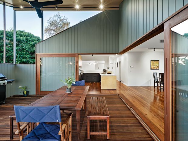 A bright, contemporary open-plan indoor-outdoor living and dining space with a timber dining table and bench, blue upholstered chairs, timber flooring and decking, and a compact kitchen beyond. Large glazed openings connect the room to leafy surroundings, while green vertical wall cladding, exposed timber framing, a pitched internal roofline and ceiling fans define the architecture.