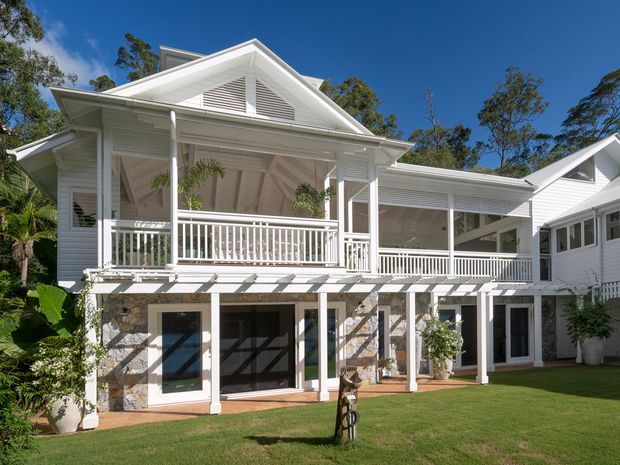Exterior view of a large white weatherboard house with a prominent covered upper-level veranda, gabled roof forms, louvred gable vents, white railings and columns, and a lower level faced in irregular natural stone. The home opens onto a manicured lawn and is surrounded by mature vegetation in bright sunlight.