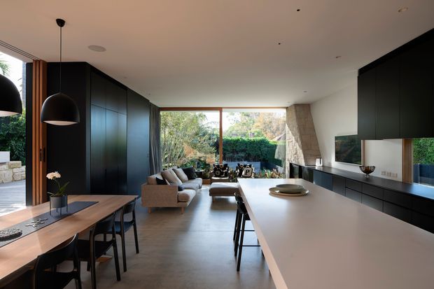 A spacious open-plan contemporary interior combining a dining area, living room and kitchen. The room features a long pale island bench with dark cabinetry, a timber dining table with black chairs, a neutral sofa, extensive glazing overlooking greenery, pendant lighting, and a distinctive angled stone-clad feature wall.