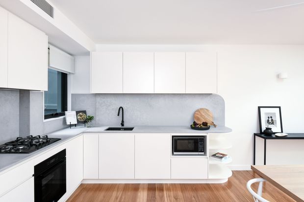 Bright contemporary white kitchen with streamlined handleless cabinetry, a grey stone-look splashback and benchtop, integrated appliances, gas cooktop, black sink and tapware, and warm timber flooring. A compact dining area and framed artwork are visible at the edge of the room.