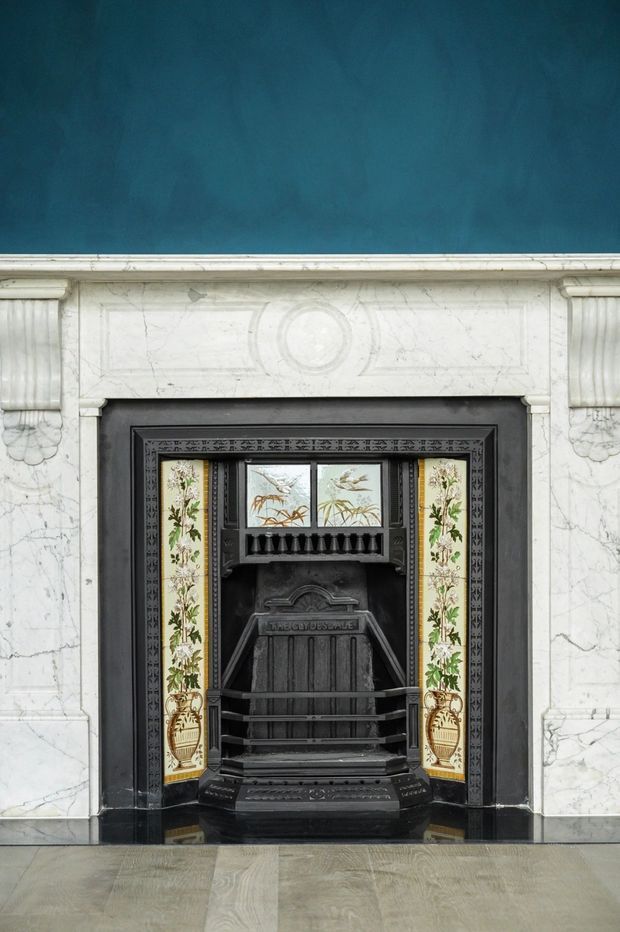 Close-up interior view of an ornate historic fireplace. The fireplace has a black cast-iron insert with decorative detailing and botanical ceramic tile panels, surrounded by a white veined marble mantel with carved classical ornament and a dark painted inner surround. A deep teal wall and pale stone or marble flooring complete the refined, heritage-style setting.