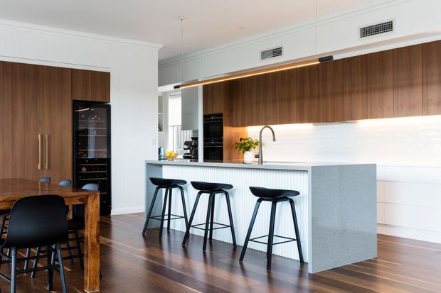 Bright contemporary open-plan kitchen and dining area with dark timber flooring, a large island with waterfall stone sides and integrated seating, white vertical fluted cabinetry, warm timber overhead cabinetry, and a white illuminated splashback. A timber dining table and black chairs sit beside the kitchen, with integrated appliances and a linear pendant light.