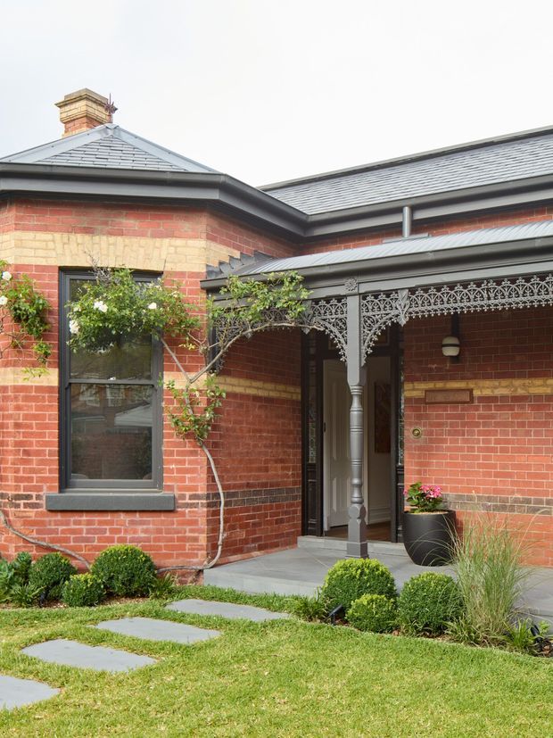 Front exterior view of a brick period house, featuring a hipped slate-tiled roof, prominent brick chimney, dark painted trim, and an ornate cast-iron verandah with decorative lacework. A paved entry path, manicured garden beds, clipped shrubs, and climbing roses frame the recessed front doorway. The image strongly suggests a Victorian-era Australian residence.