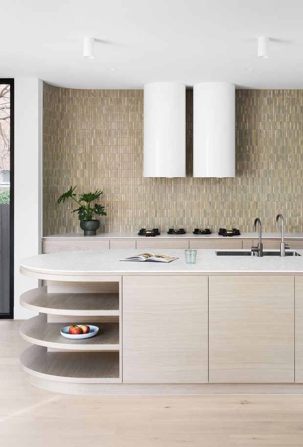 A bright contemporary kitchen centred on a large island with rounded ends and open curved shelving. The island and rear cabinetry have a pale timber finish, while the rear wall is covered in vertically arranged textured beige-grey tiles. Four black gas burners sit beneath two large white cylindrical rangehoods. A double-bowl sink with tall metal mixer taps is integrated into the island, with pale timber flooring and minimal white ceiling lighting.