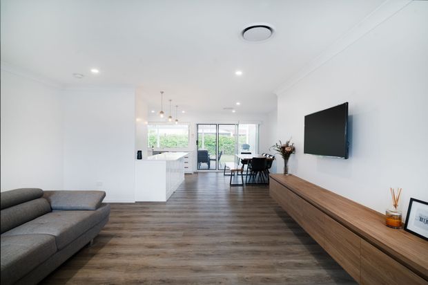 A bright, open-plan contemporary living and dining area with a white kitchen at the rear. The room features warm timber-look flooring, a large grey sectional sofa, a wall-mounted television, and a long timber media console. A compact dining table sits between the living area and kitchen, with extensive glazing opening toward greenery outside.