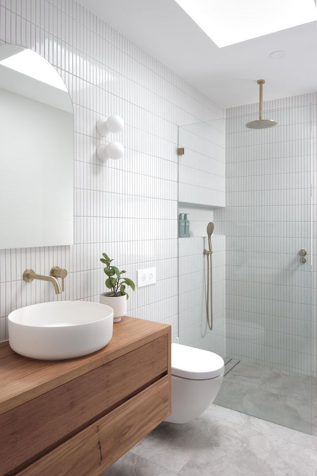 Bright contemporary bathroom with a floating mid-tone timber vanity, white vessel basin, wall-mounted brass tapware, round wall mirror, white vertically tiled walls, and a walk-in shower with matching brass fixtures. The floor is finished in light grey stone-look material, with a small potted plant adding greenery.