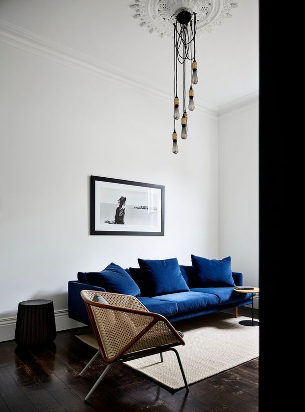 A refined living room with white walls, ornate ceiling medallion and cornice detailing, dark timber flooring, and a deep blue sofa styled with matching cushions. A sculptural hanging light with multiple exposed bulbs hangs centrally above the room. A woven cane lounge chair, black side table, framed monochrome artwork, and neutral textured rug complete the space.