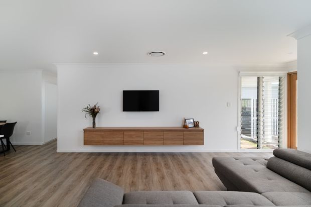 Bright contemporary living room with a large grey sectional sofa, floating mid-tone timber media console, wall-mounted television, white walls, recessed ceiling lights, and timber-look flooring. Sliding glass doors with timber framing open to an exterior area on the right.