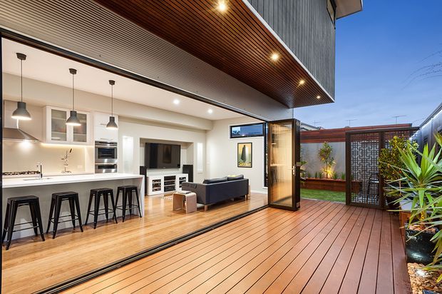 A contemporary open-plan ground-floor living area opening onto a spacious timber deck through large glazed sliding doors. The interior includes a white kitchen with island seating, integrated appliances, a dining area and a compact living room. A timber-lined soffit and ceiling, recessed lighting, and an outdoor courtyard with screening and planting are visible.