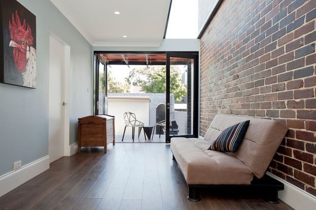 A narrow contemporary living room or sitting area with dark timber flooring, pale blue-grey walls, and an exposed red-brown brick feature wall. A low upholstered sofa sits against the brick wall, while a timber chest and artwork line the opposite wall. Full-height black-framed glazing opens to a small outdoor courtyard or terrace, with a linear skylight bringing additional daylight into the space.