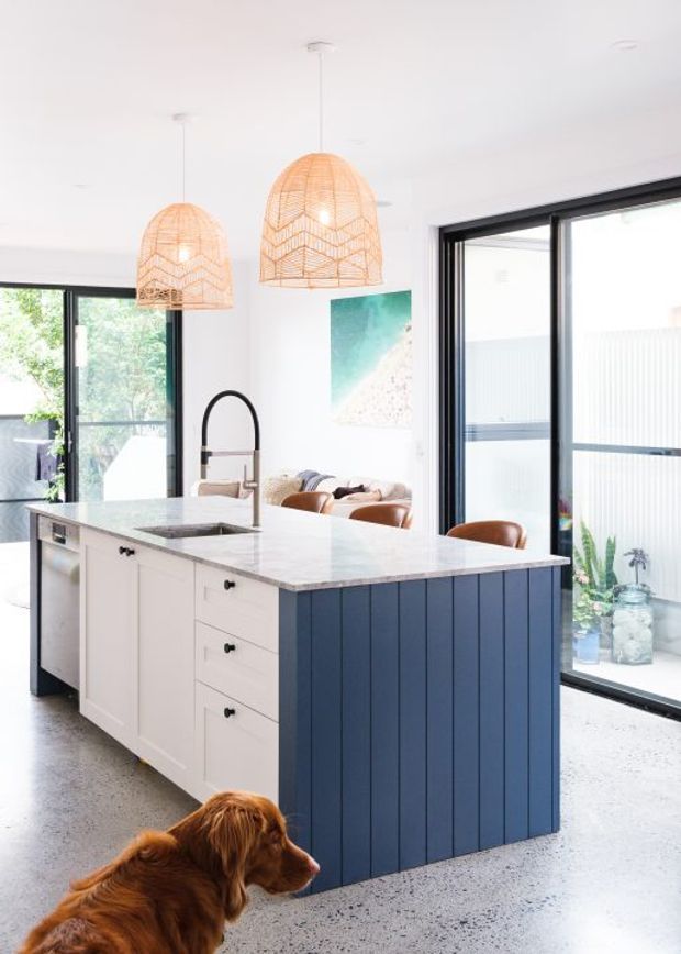 Bright open-plan kitchen centred on a large island with a white stone benchtop, white cabinetry, and a deep navy-blue vertical-panel island base. The kitchen features black-framed glazing, a gooseneck tap, integrated appliances, and two woven pendant lights. A living area is visible beyond the island, with a dog in the foreground. The space has a relaxed coastal-contemporary feel with abundant natural light.
