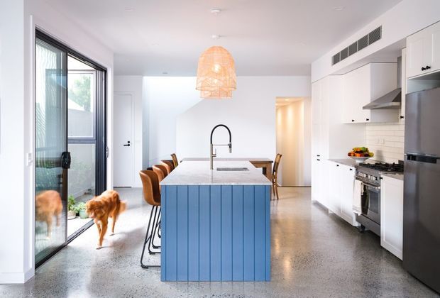 Bright contemporary open-plan kitchen with a large blue-panelled island, white cabinetry, stainless-steel appliances, pale benchtops, and polished concrete flooring. A woven pendant light hangs above the island, while full-height glazing opens to an outdoor area. The room has a clean, airy feel with warm timber bar stools and subtle natural accents.