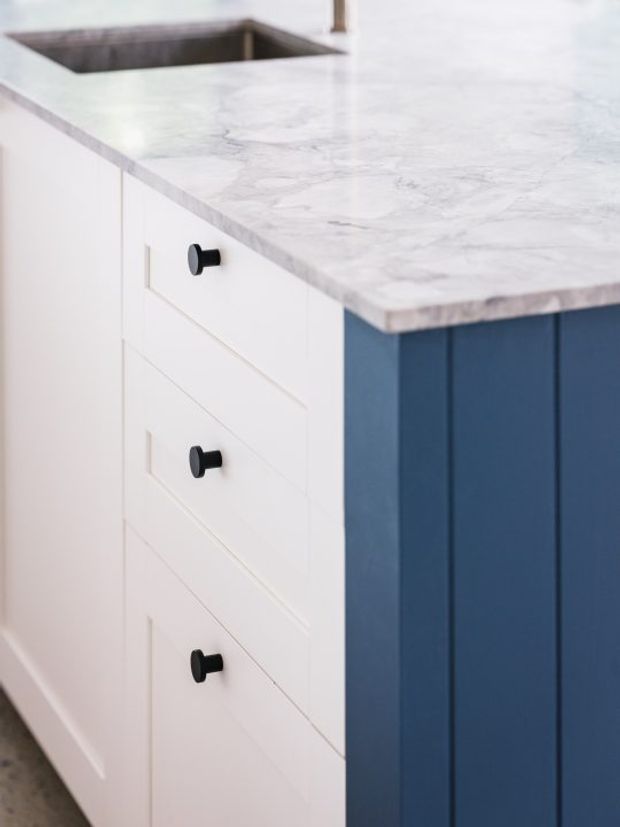 Close-up detail of a kitchen island or cabinetry featuring white shaker-style drawers with matte black knobs, a deep navy-blue paneled cabinet side, and a pale grey-white veined stone benchtop with an integrated sink.