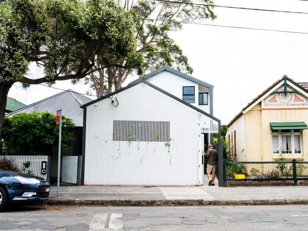 Street-facing view of a narrow contemporary infill house with a white corrugated metal facade, steep gabled roof, upper-level windows and a screened vertical opening. The modern dwelling is tightly positioned between older neighbouring houses, with mature street trees, a footpath and parked vehicle visible.