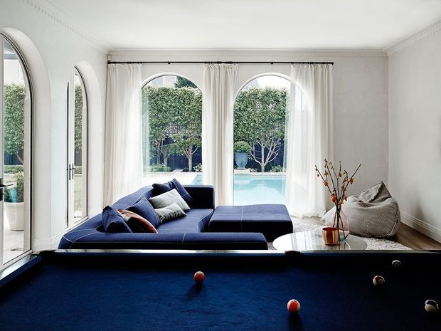 A bright, elegant living room with a deep navy sectional sofa and matching ottoman arranged beside a full-size billiards table with blue felt. Tall arched windows and glazed doors with sheer white curtains open views toward a swimming pool and landscaped garden. The room features white walls, ornate cornice detailing, dark timber flooring, a sculptural round coffee table, and understated decorative furnishings.