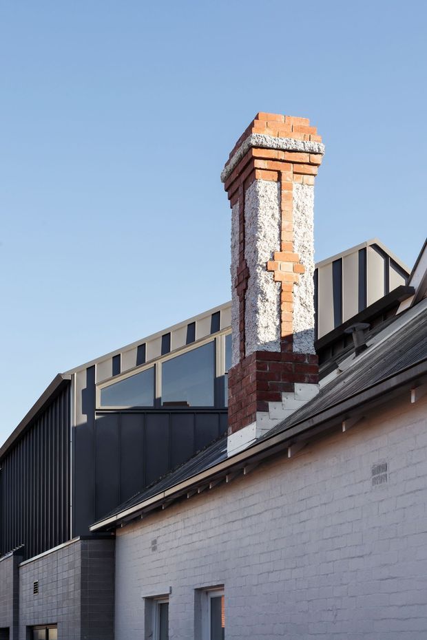 Exterior architectural photograph showing a renovated or extended masonry building with a prominent exposed red-brick chimney, white-painted brick walls, dark corrugated metal roofing, and a contemporary dark metal-clad upper structure with angular gabled forms and large glazed windows. Clear blue sky forms the background.