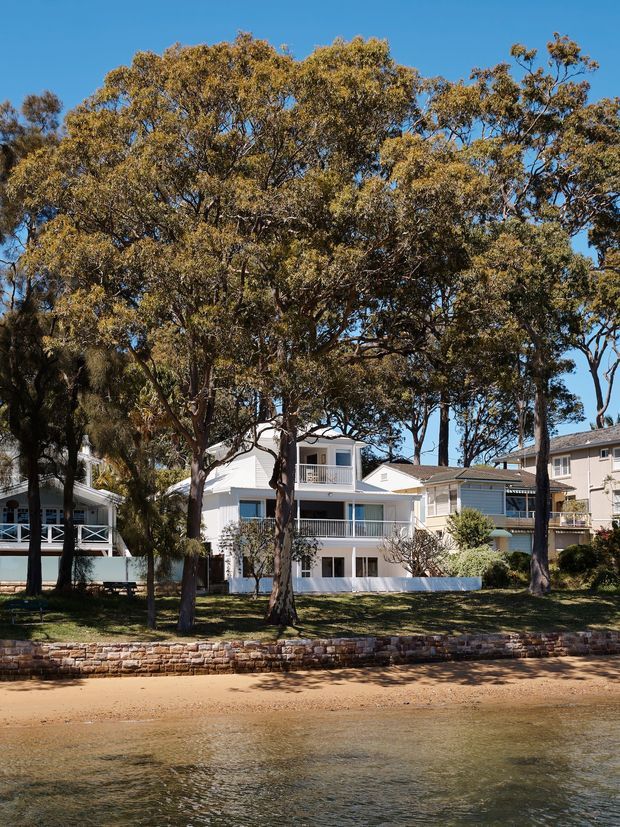A waterfront residential streetscape viewed from the water, showing a row of light-coloured multi-level coastal homes with balconies, verandas and pitched tiled roofs behind mature Australian trees. A low stone retaining wall and sandy shoreline occupy the foreground beneath a clear blue sky.