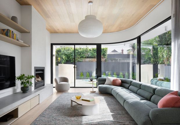 Bright contemporary living room with a large curved sectional sofa, sculptural coffee table, built-in television cabinetry and fireplace, expansive black-framed glazing, and a curved light timber-lined ceiling. The room opens visually to a landscaped courtyard and is softly furnished in muted neutral and blue-grey tones.