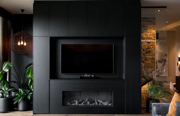 A dark, dramatic living room feature wall with full-height black built-in cabinetry framing a wall-mounted television and linear electric fireplace. Indoor tropical plants, a woven pendant light, exposed brickwork, and partial lounge seating are visible around the joinery.