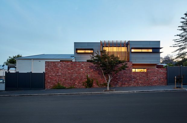 Street-facing view of a contemporary multi-level residence behind a low red-brick boundary wall and dark metal fencing. The building has a strongly horizontal composition, flat rooflines, charcoal-grey cladding, narrow illuminated windows, and a central vertical timber screen framing the upper facade. A small street tree and paved roadway occupy the foreground.