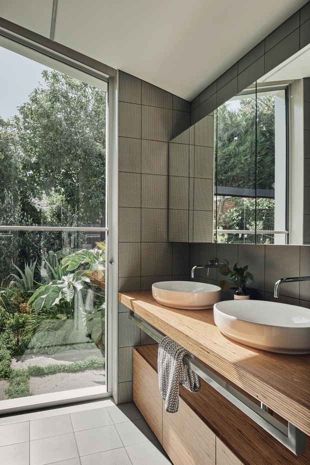 A bright contemporary bathroom with a double timber vanity, two white vessel basins, wall-mounted chrome tapware, and a large mirror. The walls are clad in dark grey small-format tiles, while large windows and a glazed door provide views to lush garden planting and abundant natural light. The vanity includes open shelving and integrated drawers, with a textured towel hanging beneath the benchtop.
