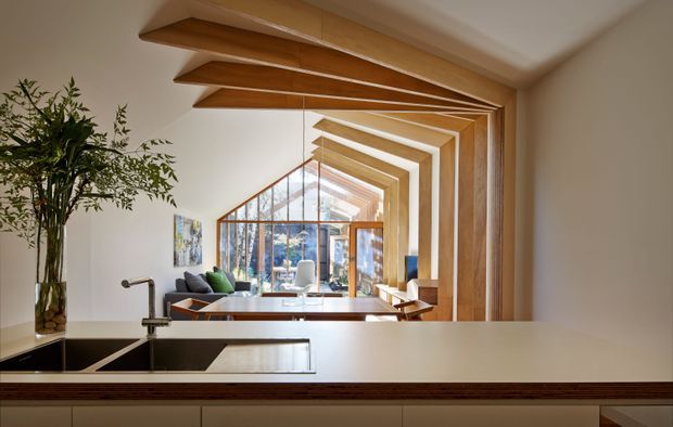 A contemporary kitchen looking through a large internal opening toward a bright living area and glazed architectural extension. The foreground has a dark undermount sink, slim metal tap, pale stone benchtop, and dark wood cabinetry. Strong repeated timber-framed ceiling and wall portals create a dramatic perspective toward the glass doors and leafy exterior. A dining or living zone with seating is visible beyond, along with a large plant and restrained modern furnishings.