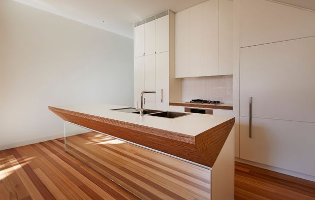 A minimalist contemporary kitchen with a sculptural island featuring a warm timber ribbed facade and white benchtop. The island includes an integrated sink and tap, while tall white cabinetry surrounds a cooktop and pale tiled splashback. Rich striped timber flooring adds warmth and contrast to the restrained neutral palette.