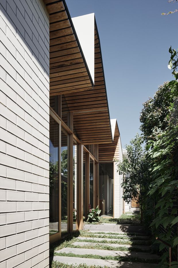 Exterior architectural view of a contemporary low-rise residence, showing a white painted brick wall, extensive timber-framed glazing, deep timber-lined roof eaves, and a stepping-stone garden path bordered by dense planting. The linear facade and repeated pitched roof forms create a sheltered, courtyard-like walkway.