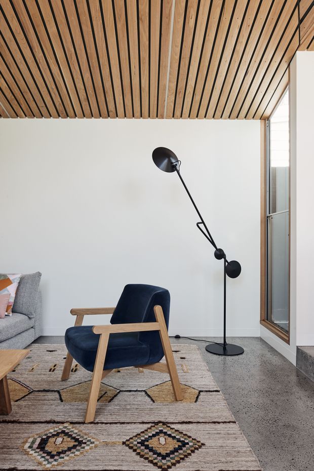 A minimalist living room corner featuring a dark blue upholstered mid-century armchair with timber arms, a grey sofa, a black articulated floor lamp, and a patterned area rug. The room has white walls, a polished concrete floor, extensive timber slat ceiling lining with black recessed gaps, and a tall timber-framed glazed opening.