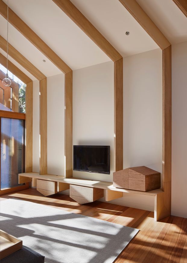 Bright, contemporary living room with a tall timber-framed feature wall incorporating a recessed fireplace, floating media cabinetry, and a low timber storage box. The room has warm timber flooring, white plastered surfaces, expansive glazing at the left, and a large pale rug illuminated by strong natural sunlight.