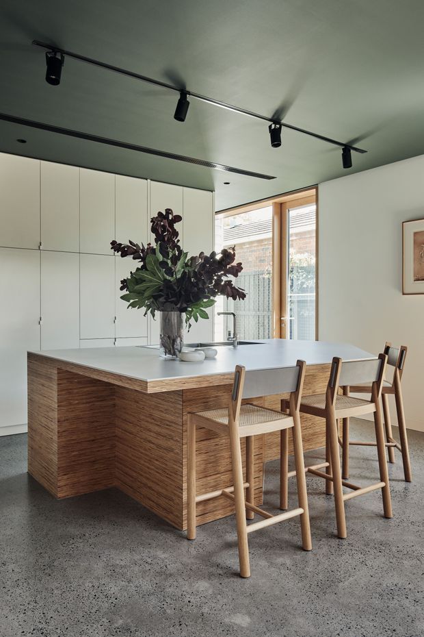 A contemporary kitchen centred on a large island with a pale solid-surface benchtop, walnut-toned timber-clad base, integrated sink and tap, and seating for four at a timber-framed breakfast bar. Full-height white cabinetry lines the rear wall, while a black track-lighting system contrasts with the pale ceiling. Timber-framed windows bring in natural light, and the space features a polished concrete floor with restrained, warm modern styling.