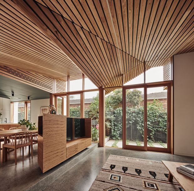 A warm, open-plan living and dining area with a dramatic slatted timber ceiling that slopes upward toward the glazed courtyard facade. Full-height timber-framed windows and doors overlook a private planted courtyard. The room features polished concrete flooring, a timber media cabinet and room divider, a dining table with chairs, and a patterned rug and coffee table. The design has a strong indoor-outdoor connection and abundant natural light.