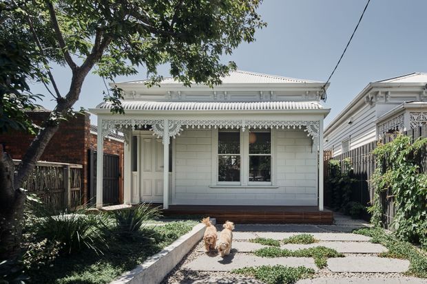Front exterior view of a compact single-storey heritage-style cottage with a pale weatherboard facade, ornate white lacework verandah, decorative gable detailing, timber door and window frames, and a paved garden path leading to the entrance. Mature planting and neighbouring period homes frame the property.