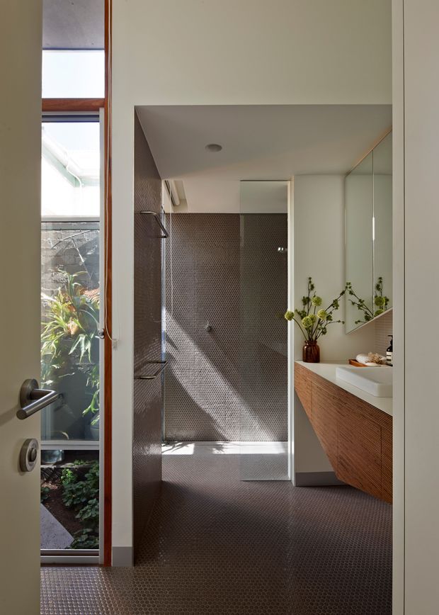 A contemporary bathroom viewed through an open doorway, featuring a walk-in shower lined in small dark mosaic tiles, matching mosaic flooring, a white vessel basin, timber-toned floating vanity cabinetry, and a large mirrored wall. A narrow glazed opening beside the door brings in daylight and views of planted greenery.
