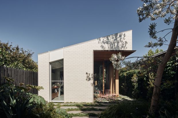 A compact contemporary single-storey residence nestled in a lush garden. The building has a crisp white masonry facade, a sharply angled mono-pitched roofline, large glazed openings, and a recessed timber-lined entry or outdoor sitting area. Stepping stones lead through dense planting to the house, creating a sheltered, natural setting.