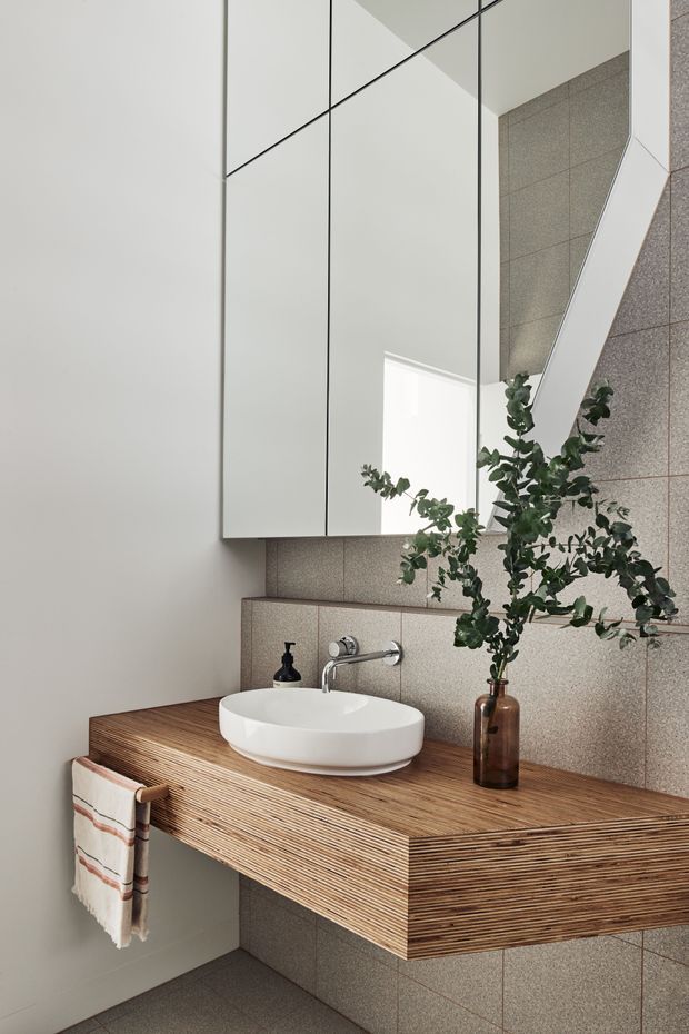 A compact contemporary bathroom vanity with a floating, horizontally laminated timber countertop and integrated side panels. A white vessel basin sits beneath a wall-mounted chrome tap, with a large mirrored cabinet above. The walls and floor are finished in small-format taupe-grey tiles, creating a restrained, natural-toned scheme. A towel rail, soap dispenser and simple botanical arrangement provide functional and decorative details.