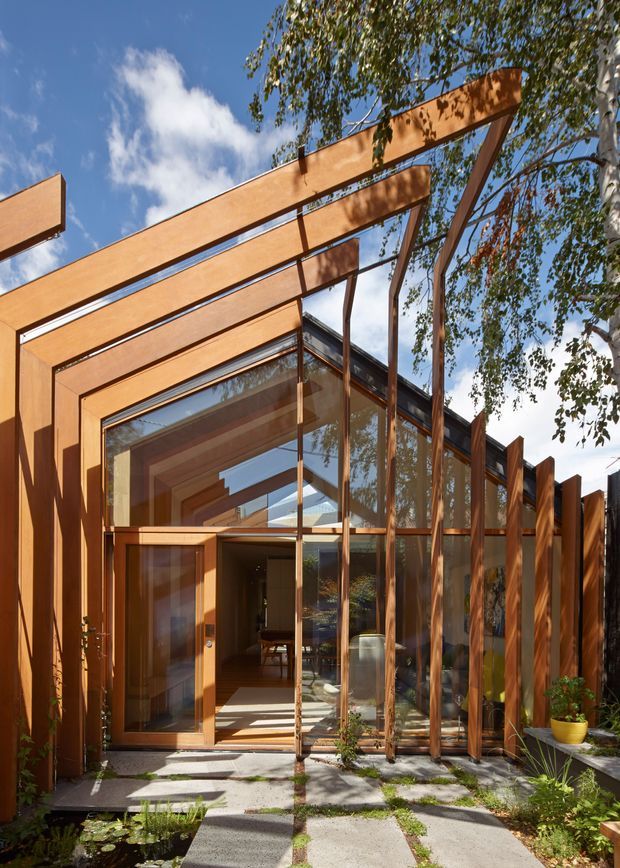 A contemporary residential entrance viewed from an outdoor garden path. Warm timber framing forms a dramatic linear pergola and vertical screen around extensive floor-to-ceiling glazing, with a timber entry door opening into a visible interior. Concrete stepping slabs, planted garden beds, and mature trees surround the facade. Strong indoor-outdoor connection, layered timber structure, and reflections in the glass are prominent.
