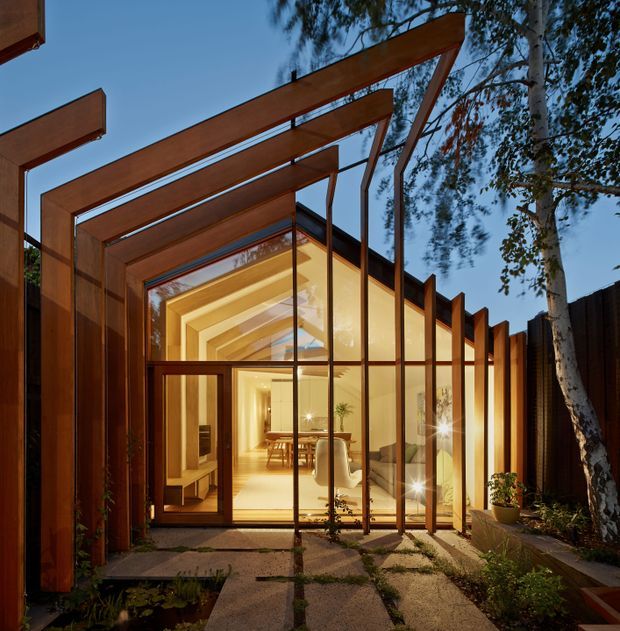 A contemporary glass-fronted residential extension or courtyard pavilion viewed from the landscaped approach at dusk. Repeated rust-toned timber or steel portal frames project outward to form a dramatic pergola-like structure, while full-height glazing reveals a warmly illuminated dining and living area inside. A patterned paved path, planting, and a mature tree frame the entrance.