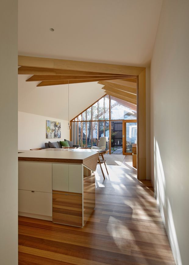 A bright open-plan interior viewed from a hallway or kitchen edge, featuring a timber kitchen island with pale cabinetry, an integrated dining and living area, extensive timber-framed glazing, and a dramatic angled ceiling with exposed timber beams or rafters. Warm timber flooring and strong sunlight create a calm, contemporary atmosphere.