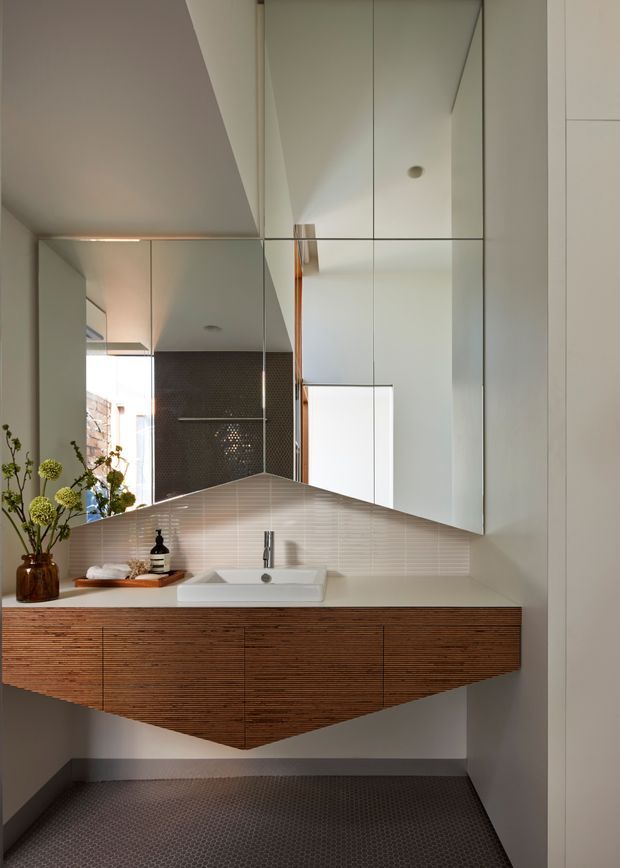 A compact contemporary bathroom featuring a wall-mounted floating vanity with richly textured horizontal timber cabinetry, a white vessel basin, chrome tapware, a large mirrored wall cabinet, and a pale linear-tile backsplash. The floor is finished in small dark mosaic tiles, while a darker tiled wall and a glimpse of an adjoining space are reflected in the mirror. A sculptural, faceted vanity profile adds visual interest.